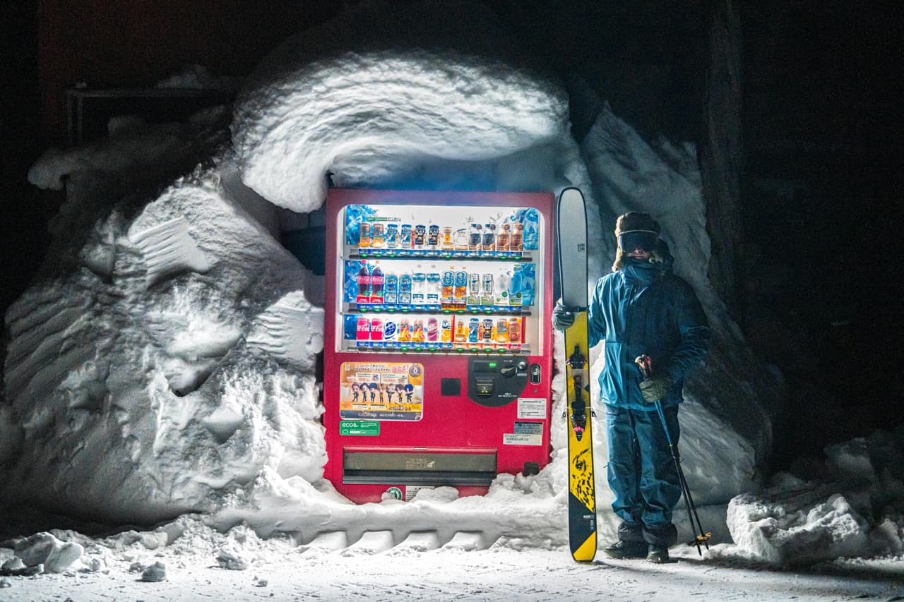 A skier beside a snow-buried vending machine at night in a Japanese ski village